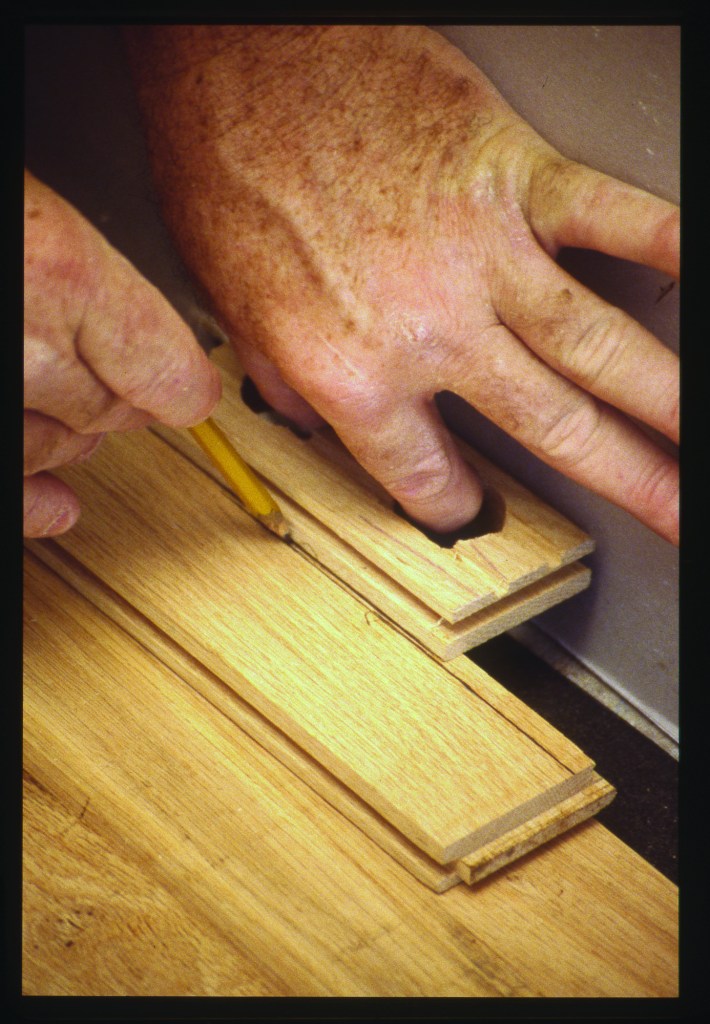 With a pencil against the block, and the tongue of the block against the drywall, scribe the entire length of the board. The tongue on the block creates expansion room for the final board. For ease of installation, cut off part of the bottom leg of the groove. After slipping the final scribed board into place, lever or wedge it tight against its neighbor and secure it with face nails.