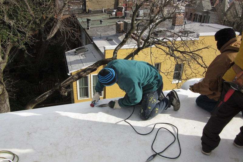 TPO single-ply membrane seams are heat-welded. Here, the installer warms up both the TPO membrane and TPO membrane flashing strip along an open roof edge using a special heat gun, which melts both pieces of a TPO roof joint. They are then pressed together with a hand roller to form a watertight seam.
