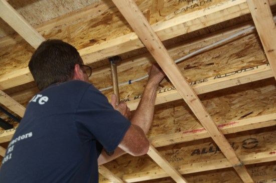 Then he reaches over and pulls the cabletight while he finishes driving the staple into the joist.