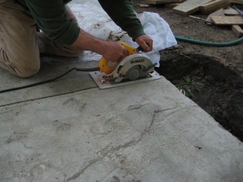 The morning after the concrete pour, the author uses a circular saw with a masonry blade to cut control joints in the concrete. These joints make it more likely that if the concrete cracks as it cures or settles in the future, it will do so in a straight line. 