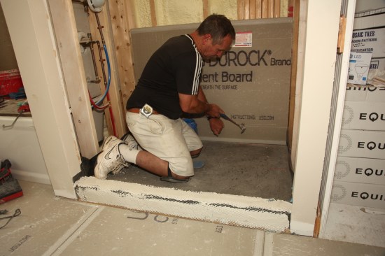 Many tile installers put the backerboard on before installing the mud base, which is acceptable if the board is kept off the surface of the pan—where it could wick up moisture and cause discoloration of the tiles. But this method requires the installer to step on the membrane, potentially exposing it to damage. Instead, wait until after the mud base is installed to install the backerboard, so that you never have to step on the membrane. With this method, the board can sit directly on top of the mud layer. Attach the board with 1 1/2 -inch galvanized roofing nails, keeping all the nails at least 2 inches above the top of the threshold.