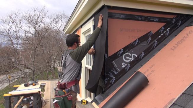 Pollard applies asphalt felt paper over the ZIP sheathing, sliding the top edge of the paper up underneath the unstapled lower edge of a strip of paper already applied to the eave under the trim board. To his left, Pollard tucks the paper under Mortairvent rainscreen fabric already installed under the window trim.