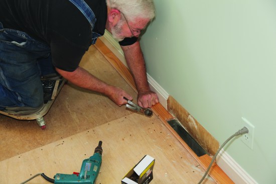 A nail spinner (in the drill on the floor) drives the flooring nails part-way. A hammer finishes driving them; each nail is set flush with the tongue.