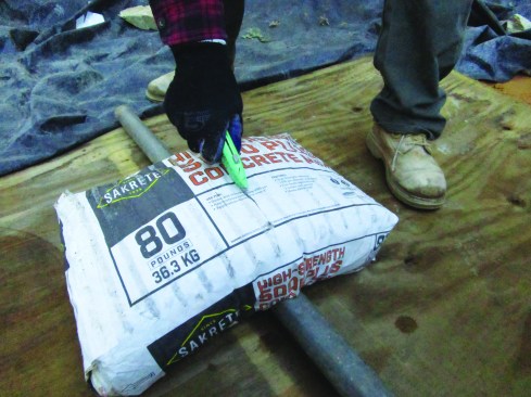 Bagged premixed concrete has many advantages and is convenient. The 80-pound bags can be arduous to handle allat once, so the author cuts the bags in half. First, he places the bag over a pipe and slits the top paper.