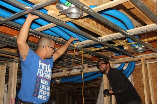 Carpenter Diego Gutierrez and project manager Travis Anderson install air supply tubing in the ceiling of a room. This photograph shows two supply registers with tubing already attached.