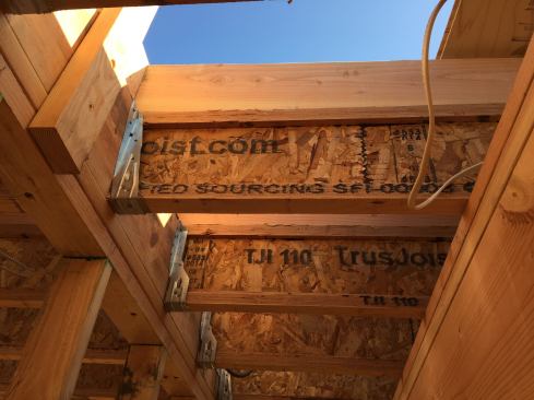 A view from below of the built-up ceiling and roof structure for the second story addition. The wood I-joist ceiling structure, viewed earlier, remains in place, with 2x framing on top of it cut to create the slope of the low-pitched roof. The joists are tied into a central girder on top of a bearing wall, using steel joist hangers.