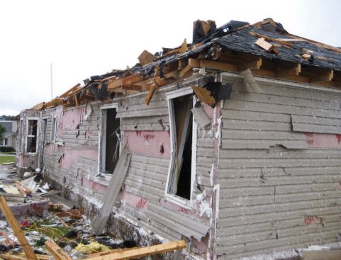 Though heavily damaged, this house may have fared better than nearby homes because it has a hip roof, which is better than a gable roof at resisting high winds. The wall seen here was knocked in by an impact that occurred between the two windows in the middle of the photo. Though the wall was sheathed with OSB — which helped hold the framing together — the connection at the top plate failed.