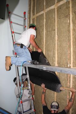 After re-insulating the wall with rock wool fiber, the crew applies sound-blocking mass-loaded sheet vinyl (
Sound Barrier HD) to the studs. Intentionally designed to be soft and massive, the vinyl weighs upwards of 120 pounds per roll. It’s a struggle for a three-man crew to maneuver the heavy rolls into position on site and install the material in the tight quarters of an existing stairwell.