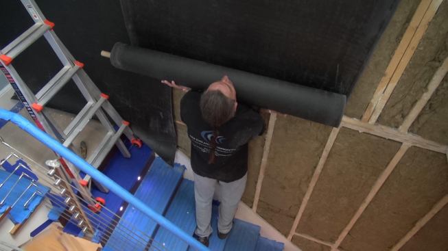 The low man has to do some heavy lifting, supporting the weight of the roll of sound-deadening sheet vinyl while the men on the staging above position and fasten the material at the top of the wall.