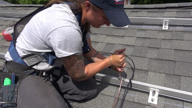 On the roof, Janarelli tightens down hardware that bonds the copper ground wire to an aluminum mounting bracket for the solar panel array. All six mounting brackets must be bonded to the grounding cable, which will run continuously back to the direct current disconnect switch and be connected to the copper grounding system for the main house.