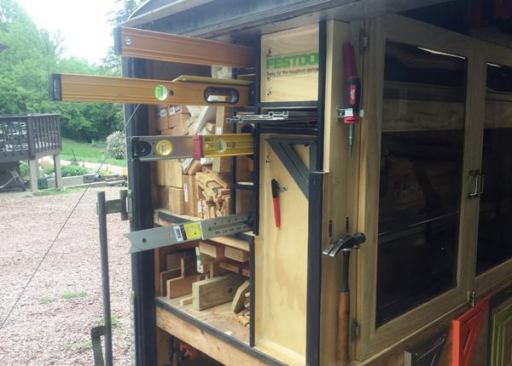 Along the left rear side of the trailer is console that is divided into cubbies. The large upper cubby at the side of the trailer can hold lumber up to 8 feet long. The cubby below that is only 5 feet long and is used to store scrap. The smaller inboard cubbies house 4-, 6-, and 8-foot levels, along with an adjustable 3/4/5 square. Note the horizontally oriented cubby just below the Festool sign. Built to house track saw rails, it is a hollow box that passes through a cabinet. From the inside of the cabinet it looks like an overly thick shelf.