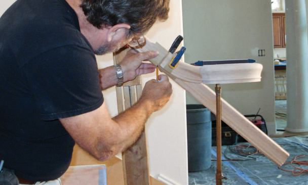 With the up-easing cut to the rake angle, the author plumbs the volute down to the centerline of the starter step, clamps the fitting in place, and scribes the cut on the handrail.