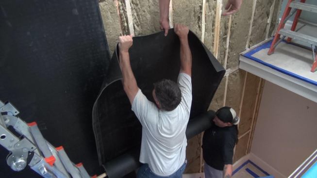 The crew unrolls the mass-loaded sheet vinyl vertically, working along the wall. Joints are positioned between studs (within the stud bays), so that overlaps won’t create a bulge in the wall when drywall is applied.