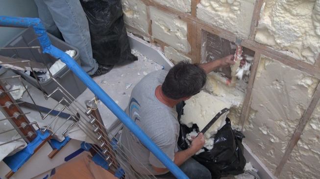 Working carefully to avoid damaging an existing water pipe in the wall, the crew removes low-density, open-cell, spray-applied polyurethane insulation from the existing party wall. Open-cell foam transmits sound less readily than high-density closed-cell spray foam, but rock wool fiber performs better than either, so this wall will be re-insulated with special sound-absorbing rock wool.