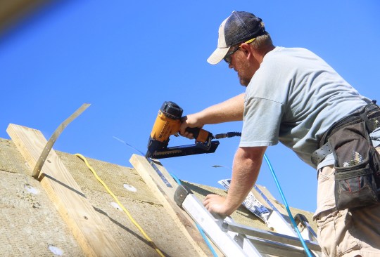 A framer nails steel straps onto the 2x6 roof strapping, to connect the strapping across the roof peak to strapping on the opposite side of the roof.