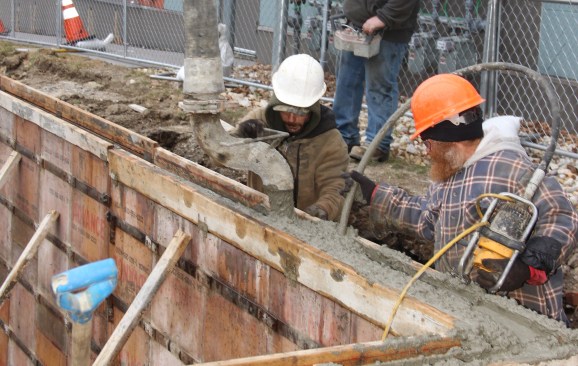 While the pump truck operator controls the flow of concrete with a remote control, concrete masons direct the mix into the forms and consolidate the concrete with a mechanical vibrator.