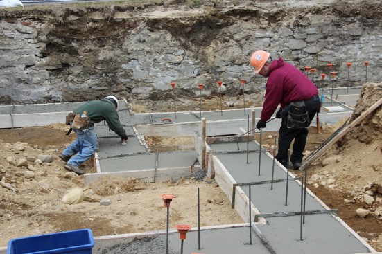 Concrete masons finish trowel the freshly poured footing and hook bent half-inch rebar under the existing rebar in the footing at pre-marked intervals.